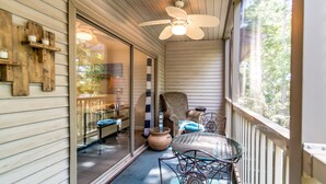 Une véranda couverte avec un ventilateur de plafond, une chaise en osier, une table avec des chaises et une vue sur la verdure à travers la porte vitrée.