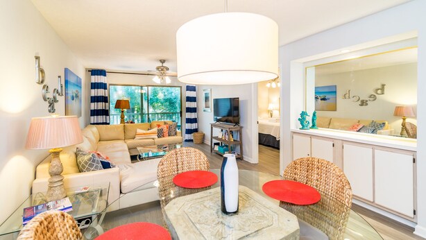 Un salon moderne avec un canapé, une table basse et des fauteuils en osier. On y trouve également un téléviseur et un coin repas avec un buffet blanc.