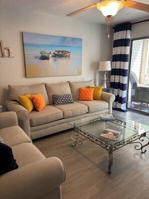 Un salón con un sofá color beige, cojines estampados, una mesa de centro de cristal y un cuadro de un barco en la pared.