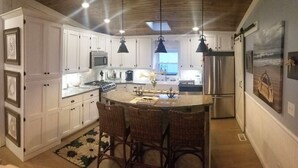 Una cocina con armarios blancos, una isla central con taburetes de bar, iluminación colgante y vistas al exterior.