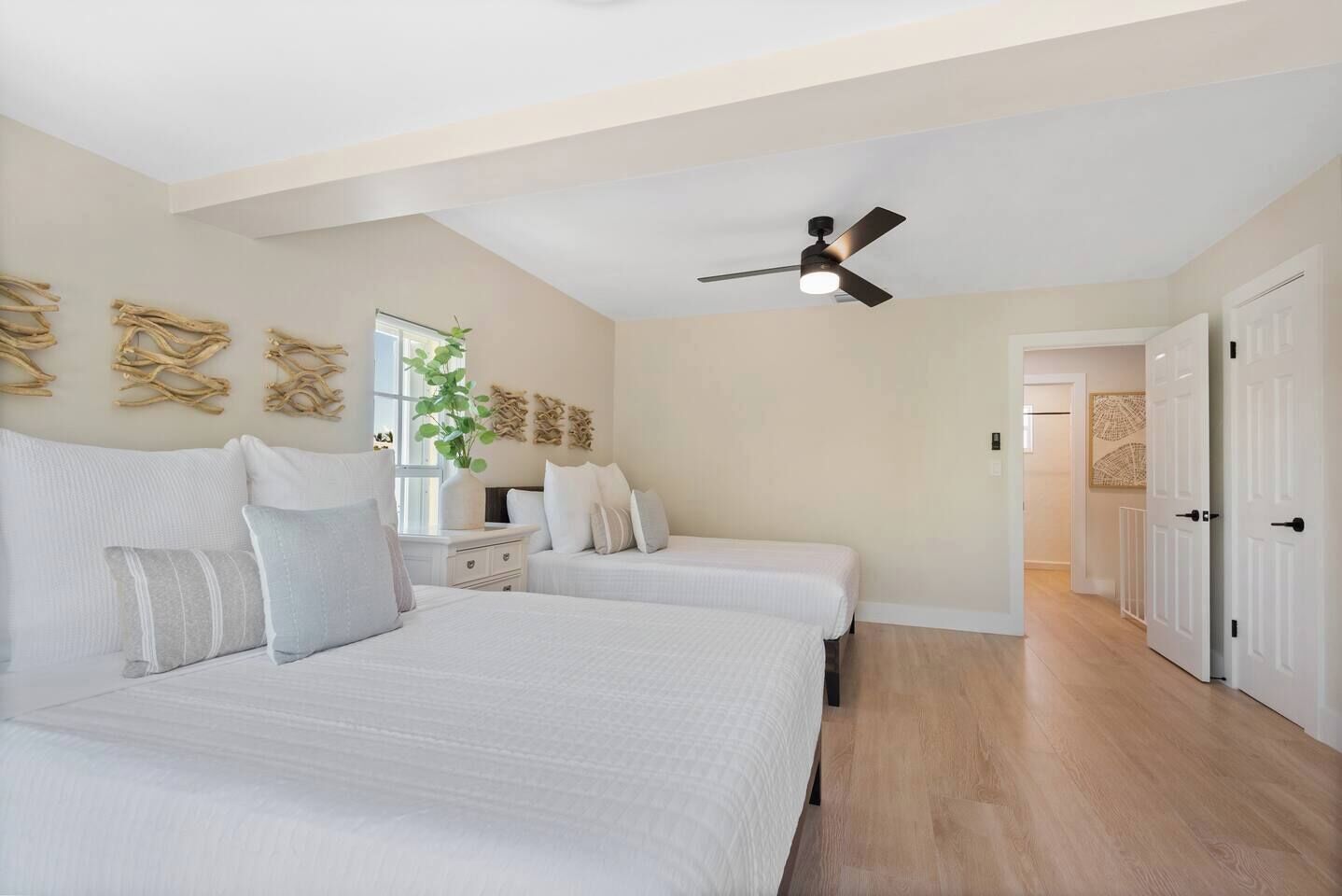 A bedroom with two beds, a ceiling fan, a window with a plant, and a hallway leading to a door.