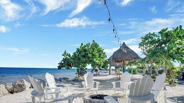 A beach setting with white chairs, a thatched-roof structure, and a fire pit. - Casa Largo (Key Largo)