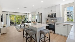 A modern kitchen with white cabinetry, a central island with stools, a dining area with a view, and a large sliding glass door. - Casa Largo (Key Largo)