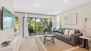 A modern living room with a grey sofa, a coffee table, and a view through glass doors to an outdoor area with chairs and plants. - Casa Largo (Key Largo)