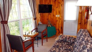 A cozy living room with a wooden interior, a television mounted on the wall, a sofa, two chairs, and a small table with chess pieces. - Lakeview cabin on stunning Lake Vermilion (Tower)