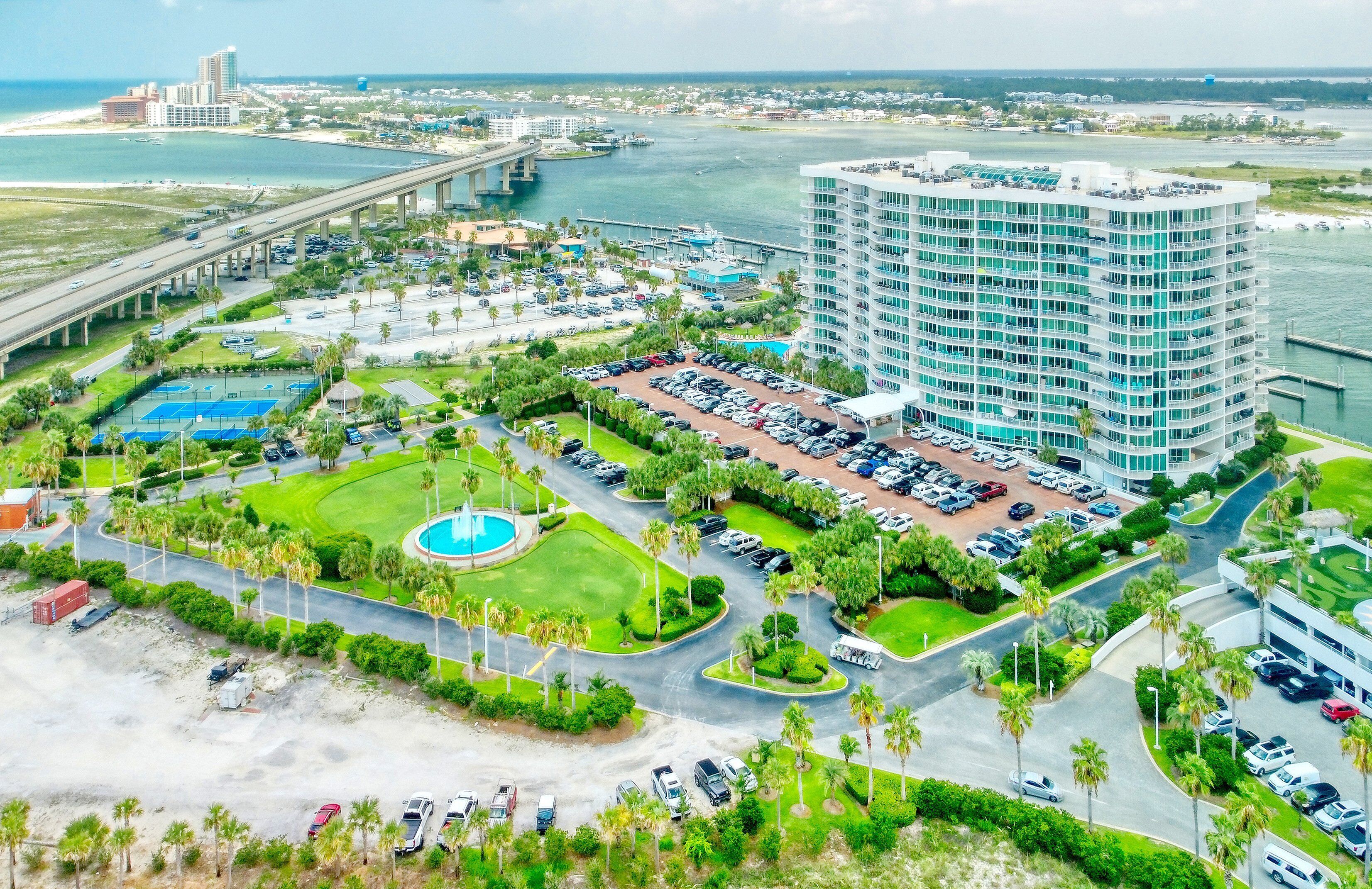 A high-rise hotel with a swimming pool, parking lot, and a view of the ocean and bridge.