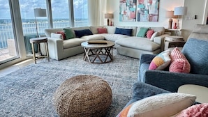 A modern living room with a large window, a sectional sofa, a round coffee table, and a blue armchair. - Renovated Caribe Penthouse 3/3 Sleeps 12 (Orange Beach)