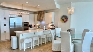 A modern kitchen with white cabinetry, a stainless steel refrigerator, a central island with bar stools, and a dining area with white chairs. - Renovated Caribe Penthouse 3/3 Sleeps 12 (Orange Beach)