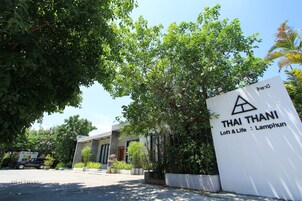 Letrero de Thai Thani Loft & Life Lamphun, un edificio moderno con fachada de concreto y amplias ventanas, rodeado de exuberante vegetación.
