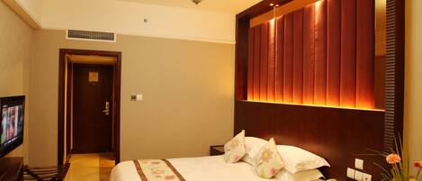 A hotel room with a large bed, a television on a stand, a bedside table with a phone and a vase of flowers, and a wall-mounted light fixture.