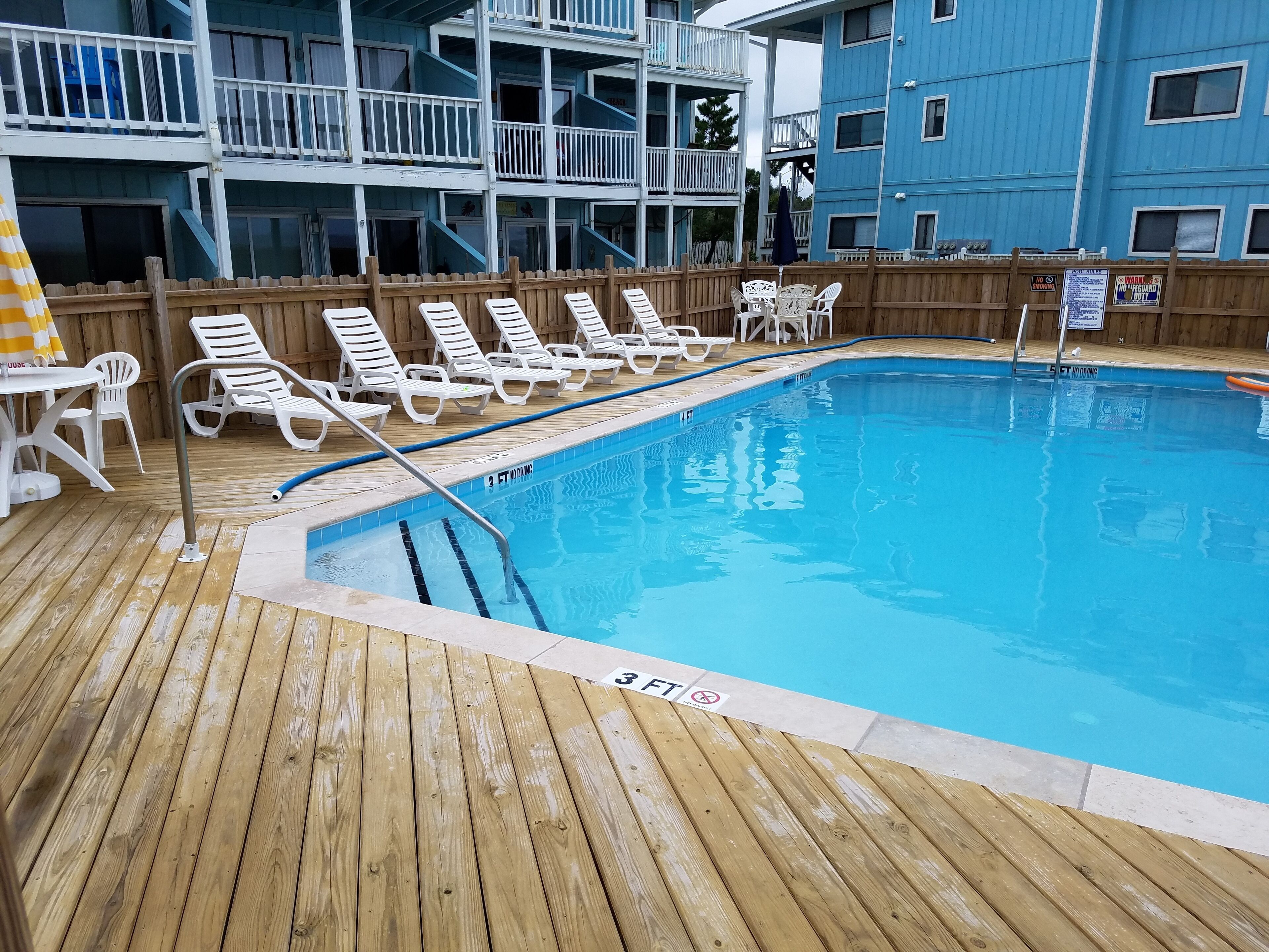A pool area with wooden deck, white lounge chairs, and a blue building in the background.
