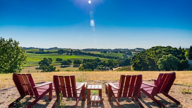 Un área de estar al aire libre con encanto, con sillas y mesas de madera, que ofrece vistas a un valle exuberante y colinas lejanas bajo un cielo azul despejado.