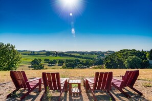 Un área de estar al aire libre con encanto, con sillas y mesas de madera, que ofrece vistas a un valle exuberante y colinas lejanas bajo un cielo azul despejado.
