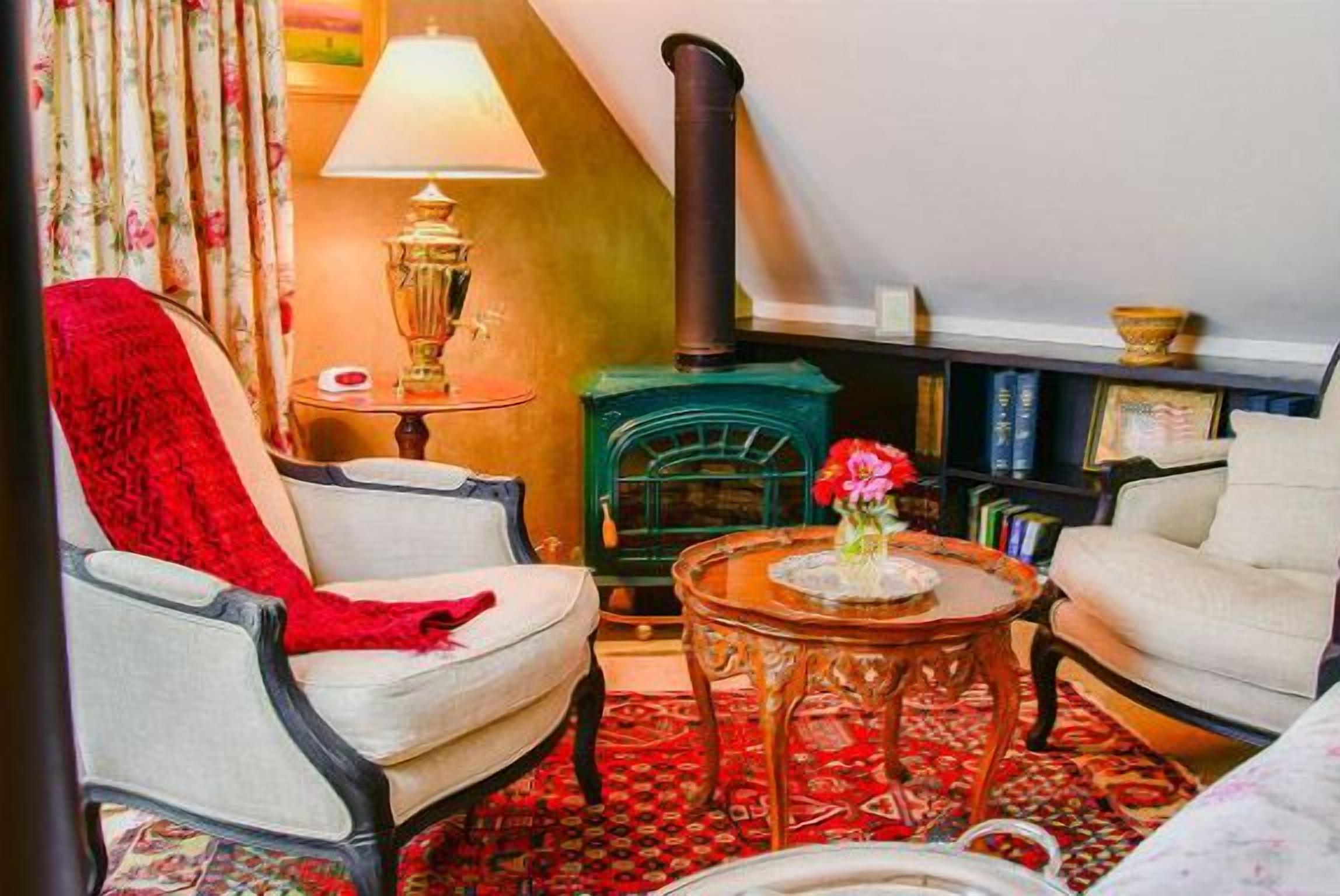A cozy living room with a fireplace, a red throw blanket on a chair, a lamp, and a bookshelf.