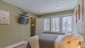 A bedroom with a bed, a TV mounted on the wall, a closet, and a window with a view of trees.