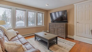 A living room with a brown leather sofa, a coffee table, a TV on a cabinet, and large windows.