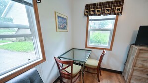 A small room with a glass-top table, two wooden chairs, a blue couch, a wooden dresser, a window with a plaid valance, and a framed picture on the wall. - Two Bed Two Bathroom Cabin with kitchenette area (Williams Bay)