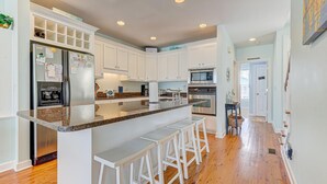 A modern kitchen with white cabinets, granite countertops, a stainless steel refrigerator, a built-in oven, and a microwave. There are three white bar stools at the island. - 🌊Mission SEA Accomplished🌊 Ocean Views, Private Grill, Steps 2 Beach (Kure Beach)