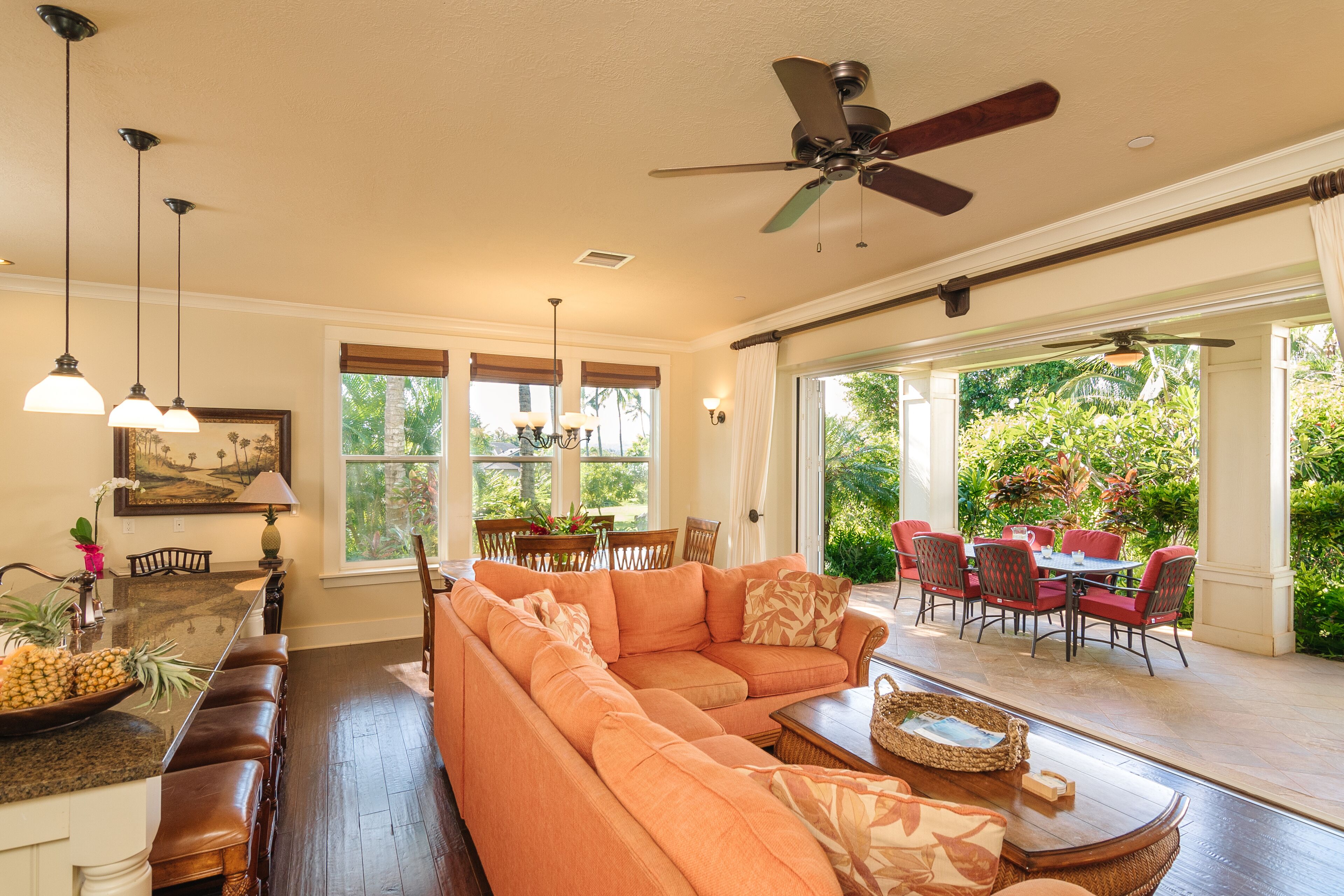 A spacious living area with a large orange sofa, dining table, and chairs. There are ceiling fans, pendant lights, and large windows offering a view of the outdoors.