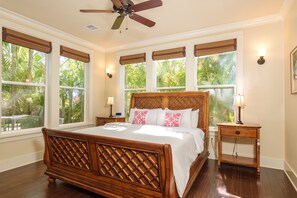 A bedroom with a wooden bed, large windows with blinds, a ceiling fan, and bedside lamps.