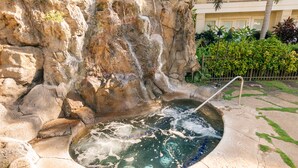 A hot tub with a waterfall feature, surrounded by a stone wall and lush greenery.