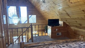 Eine Holzhütte mit Schlafzimmer im Erkerbereich, einer Treppe, einer Chandelierleuchtre und einem Fernseher auf einer Kommode.