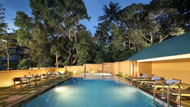 A swimming pool with lounge chairs and tables, surrounded by trees and a building with a green roof. - PepperVine (Peermade)