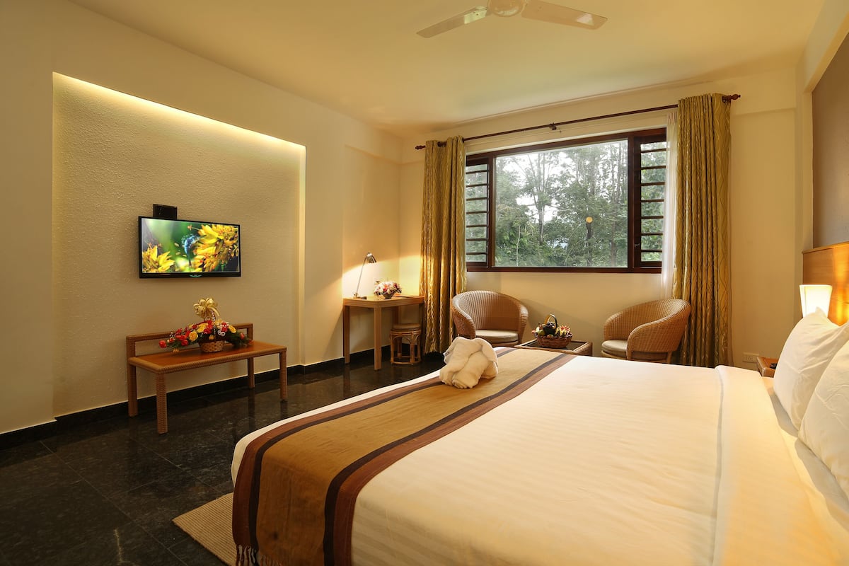 Quarto de hotel com uma cama grande, televisão, escrivaninha com luminária, duas cadeiras de vime e janela com cortinas.