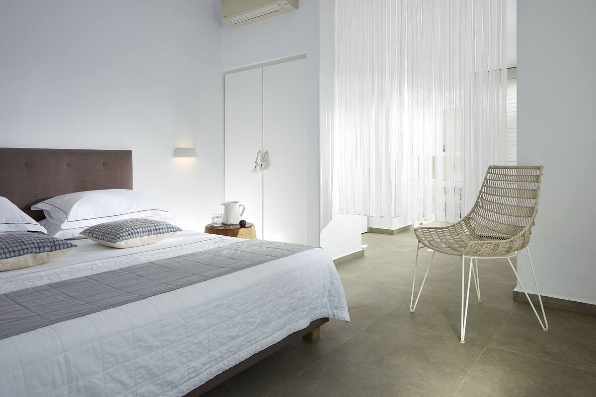 Ein Schlafzimmer mit einem Bett, Kissen, einem Sessel, einem Nachttisch und einem Fenster mit Vorhängen.