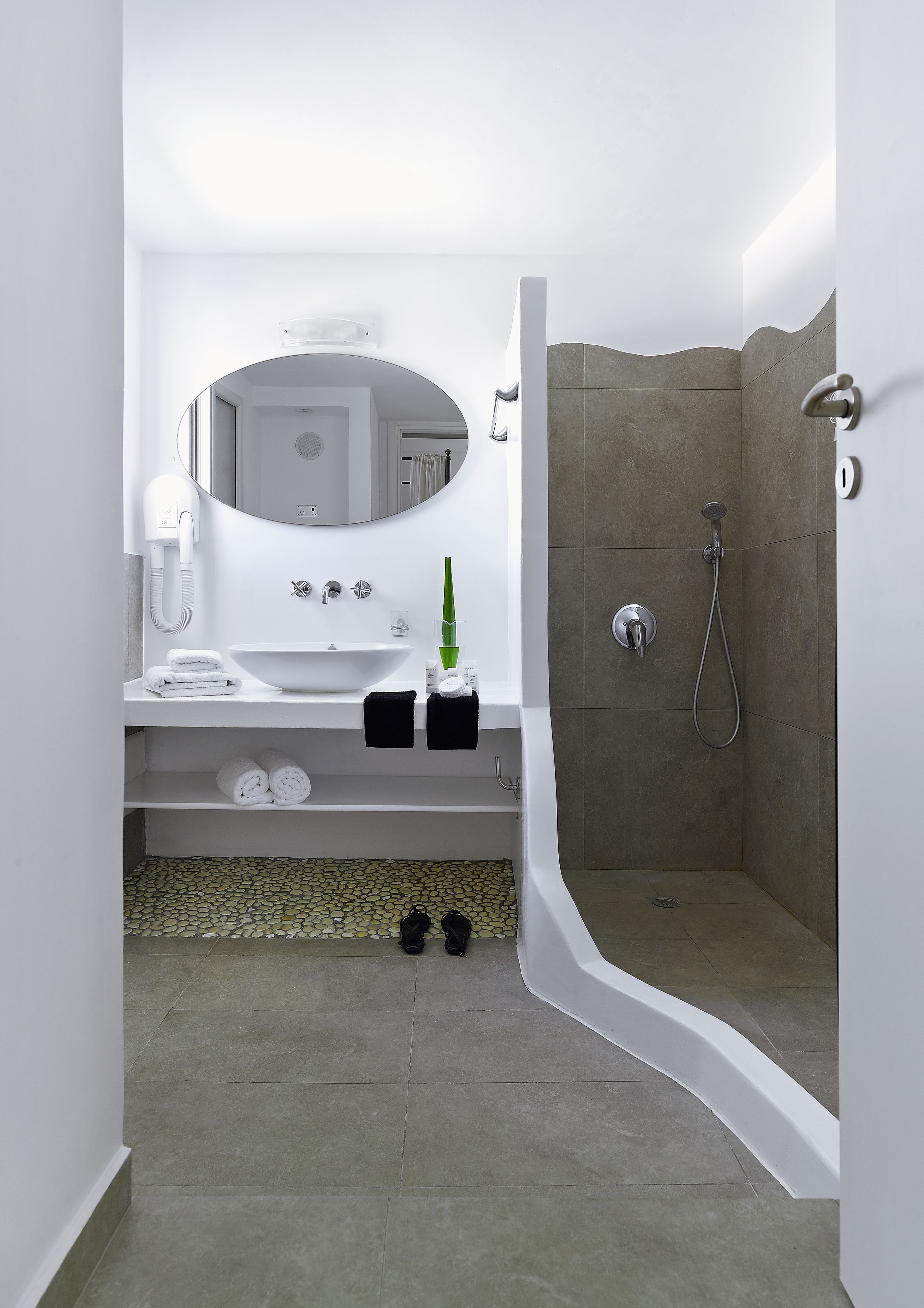 Un baño moderno con tocador blanco, un espejo grande, ducha con cabezal de ducha manual y piso de azulejos.
