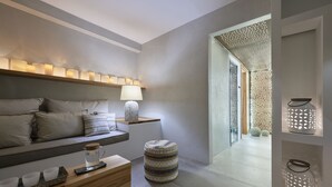 A modern, minimalist room with a sofa, a wooden side table with a lamp, a small ottoman, and a tray with cups and a pitcher. - Santa Maria Luxury Suites & Spa (Milos)