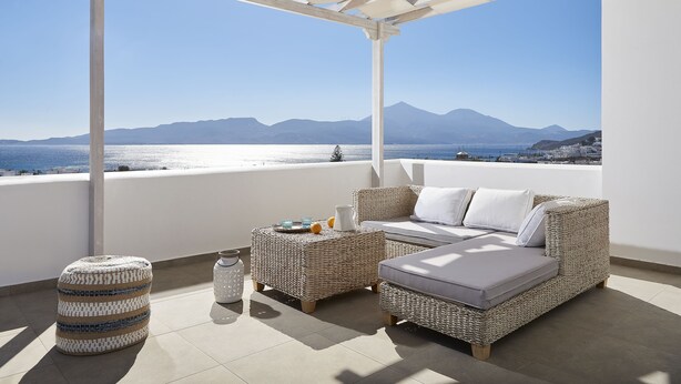 A balcony with wicker furniture, a glass of water, and a view of the sea and mountains. - Santa Maria Luxury Suites & Spa (Milos)