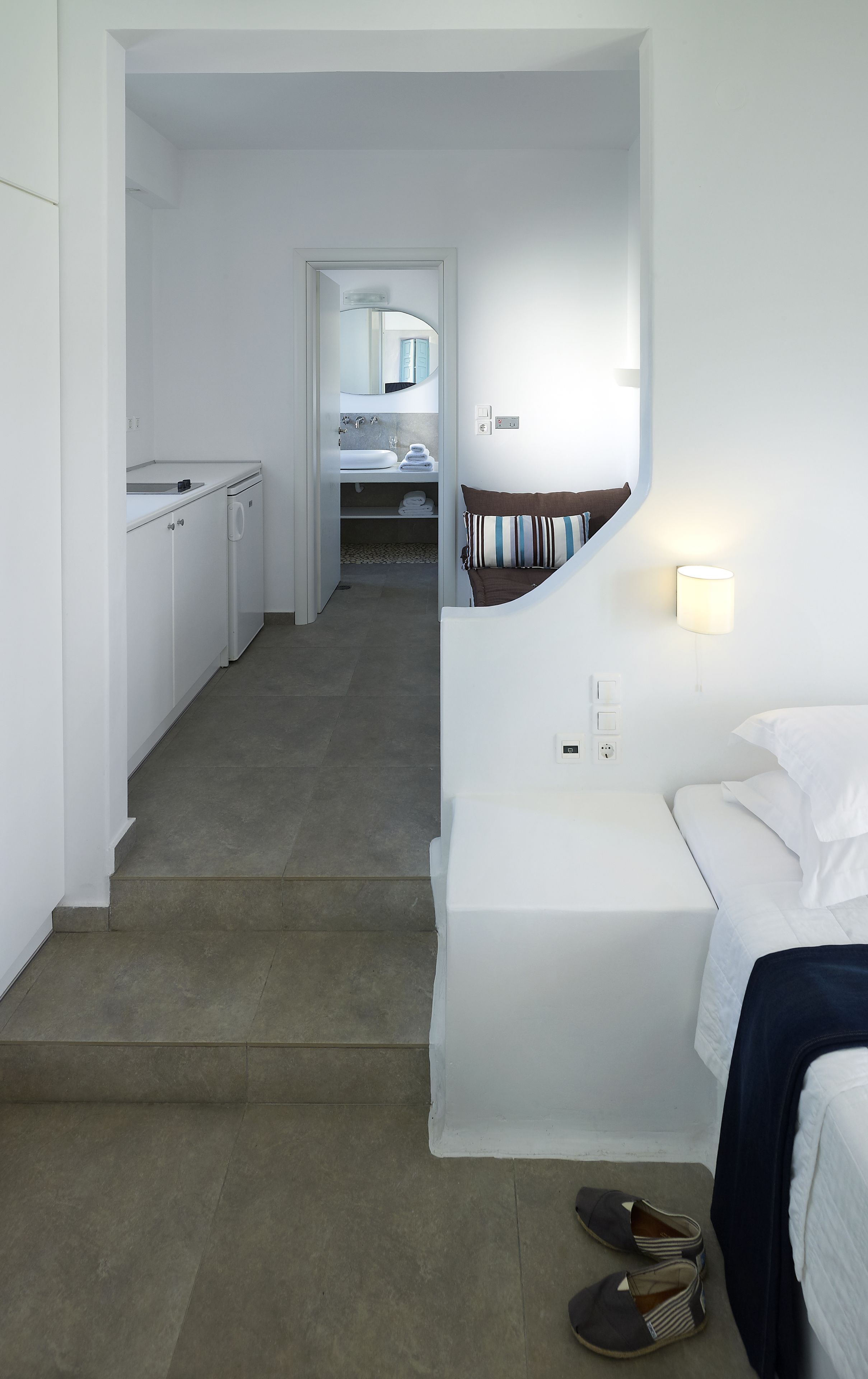 Een hotelkamer met een wit bed, een klein nachtkastje met een lamp, uitzicht op de badkamer en een gang naar de keuken.