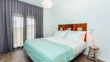 A neatly made bed with a teal bedspread, a wooden headboard, and a small wooden stool with a glass beside it.