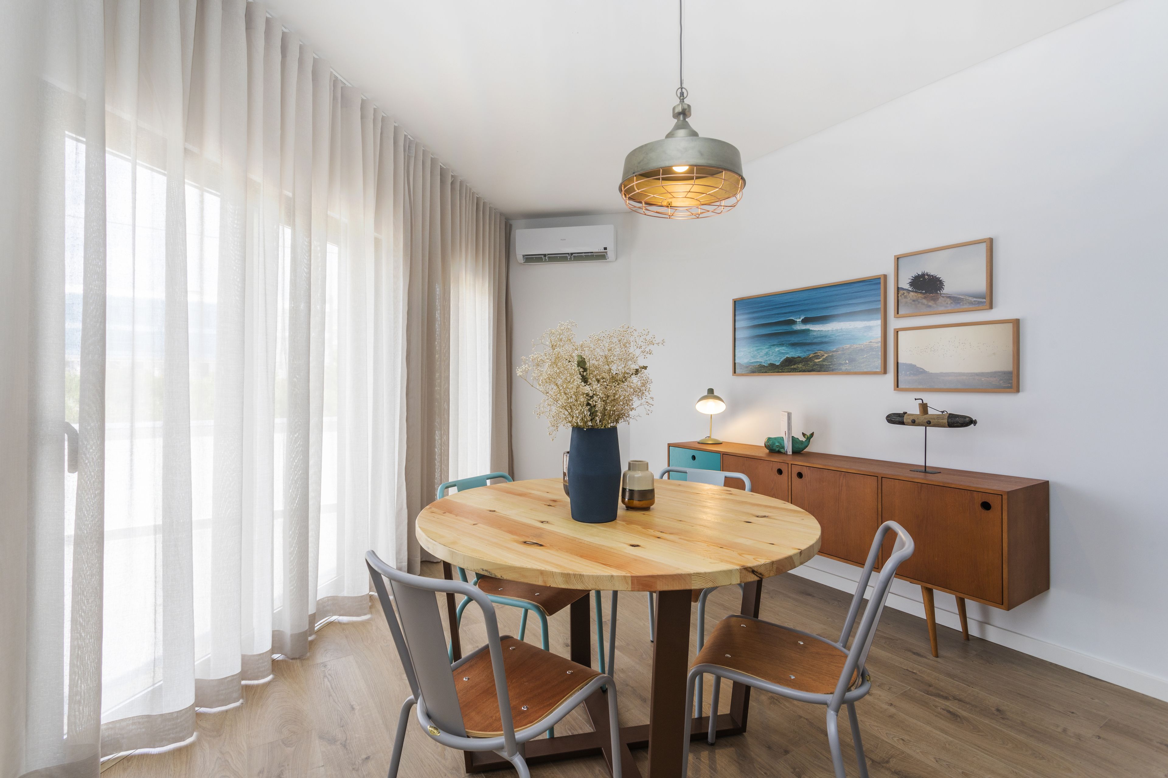 A dining area with a round wooden table, four chairs, a vase with flowers, a pendant light, and a wall-mounted air conditioner.
