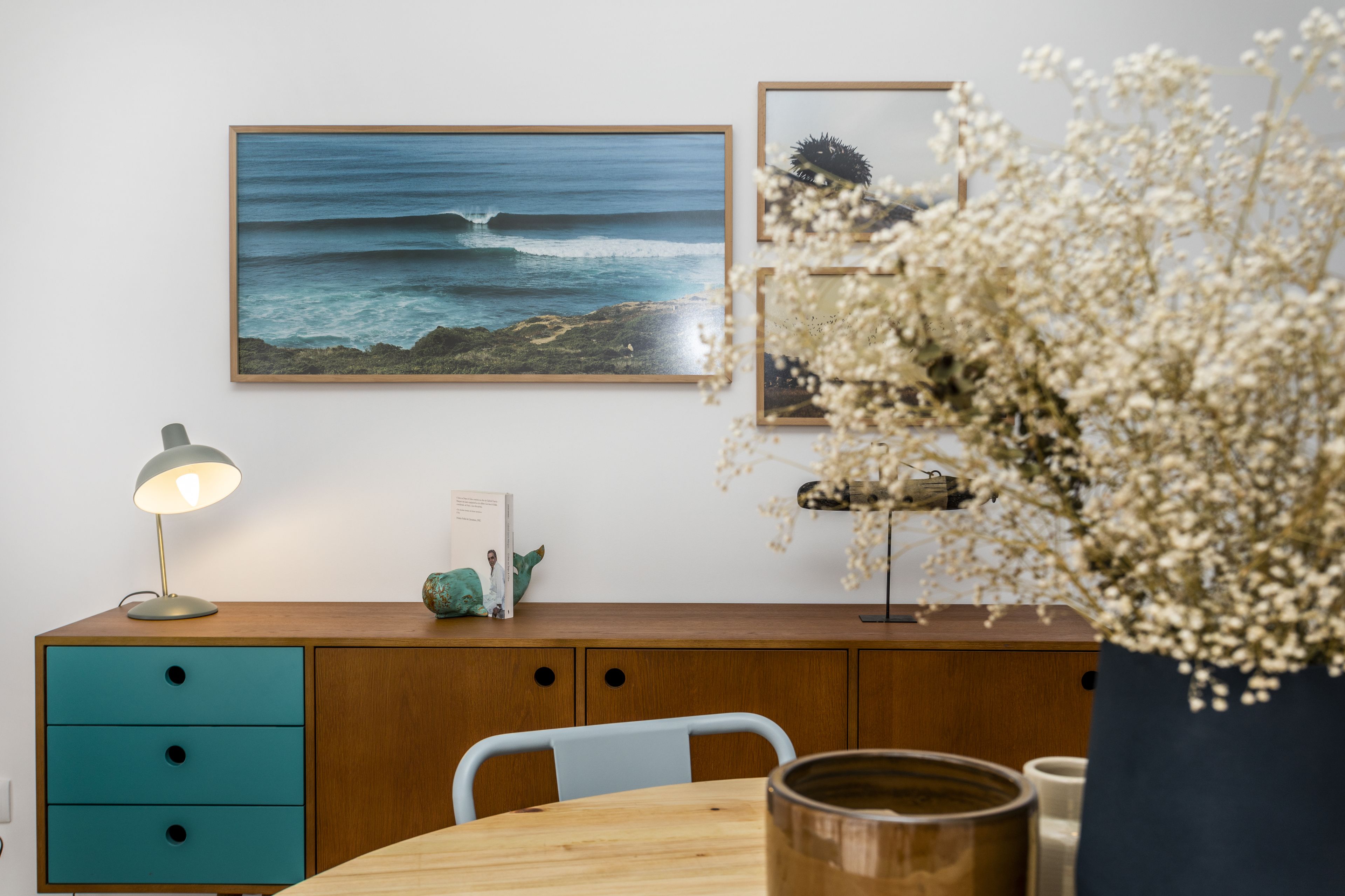 A wooden desk with a teal drawer unit, a framed ocean landscape, a small potted plant, and a vase with white flowers.