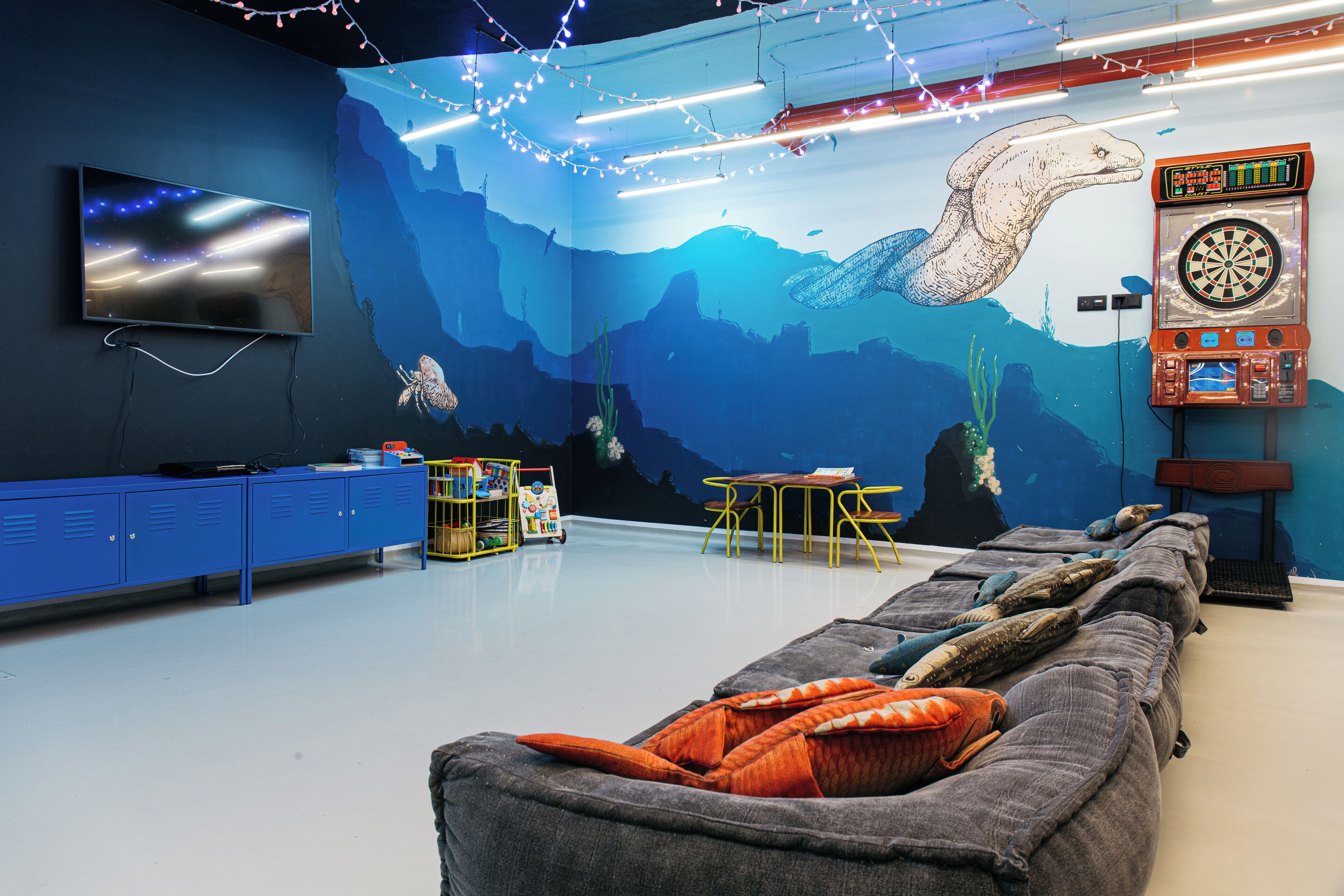 Een kamer met een groot muurschildering van een zeemonster, een flatscreen televisie, een dartbord en een bank met kussens.