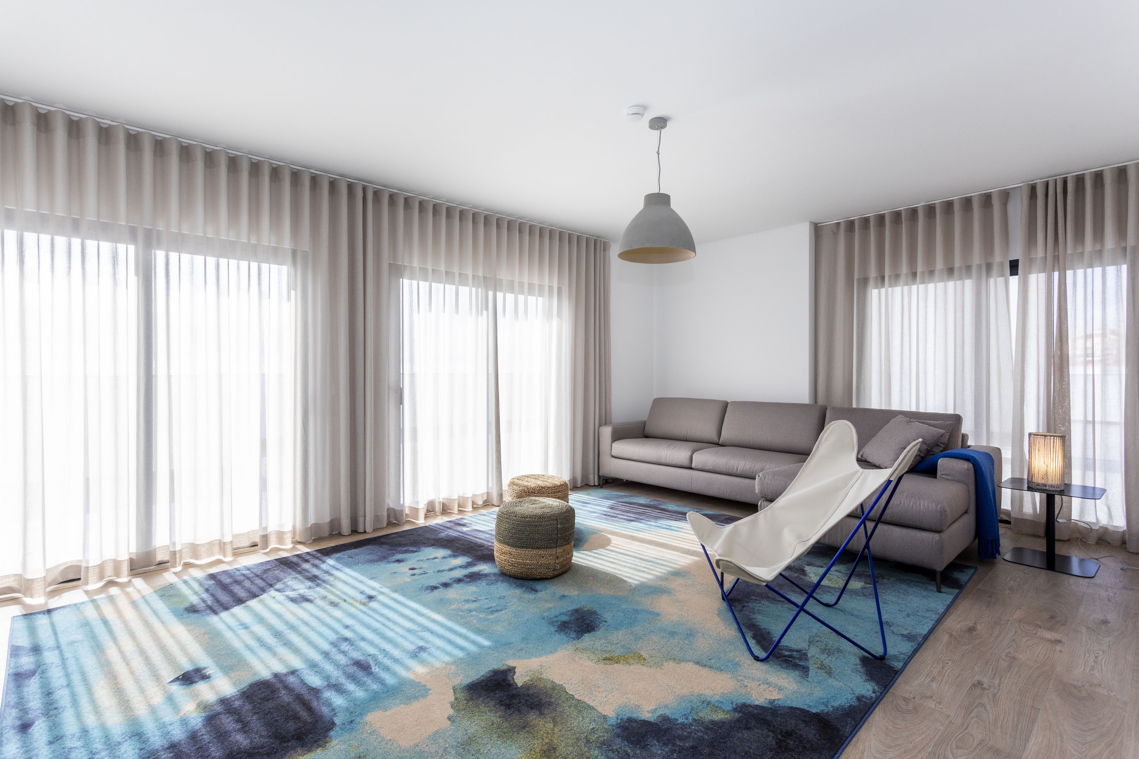 Un salon moderne avec un canapé, un pouf et un fauteuil, de grandes fenêtres avec des rideaux légers et un tapis à motifs.