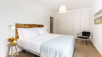 A bedroom with a wooden headboard, white bedding, a light blue throw blanket, a small wooden stool with a vase of flowers, a black chair, and a white wardrobe.