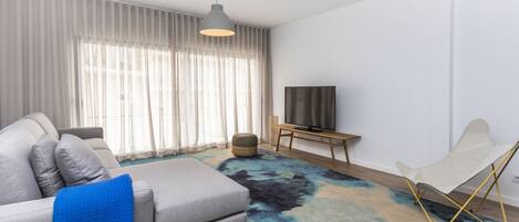 A modern living room with a grey sofa, a blue throw blanket, a black and blue rug, a television on a wooden stand, and a white folding chair.