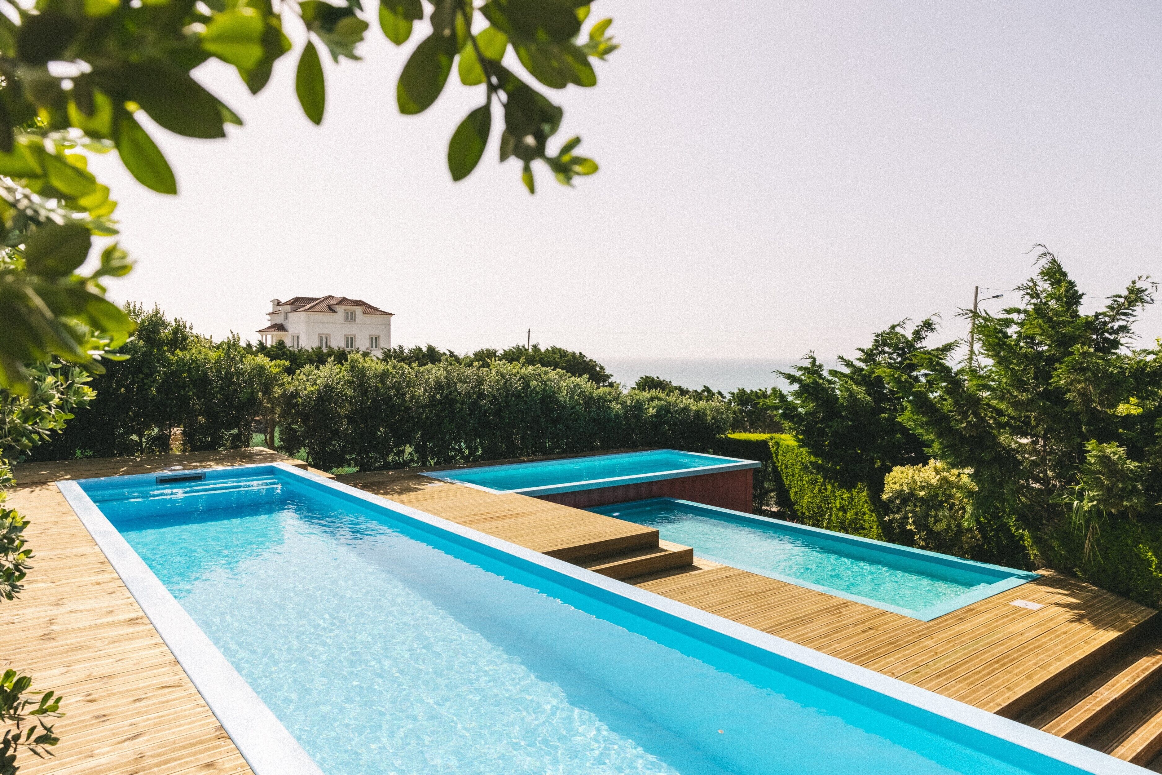 A rectangular swimming pool with wooden deck edges, surrounded by lush greenery and a view of the sea.