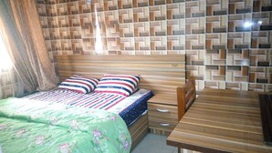 Ein Schlafzimmer mit einem hölzernen Kopfteil, einem Bett mit gestreifter Bettwäsche, einer grün-blumigen Tagesdecke und einem Nachttisch aus Holz.