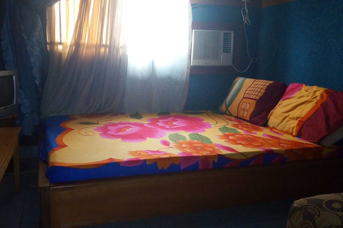 Une chambre avec un lit, une petite table, un téléviseur et une fenêtre avec des rideaux.