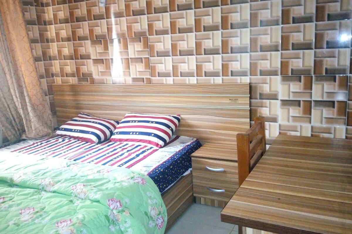 Une chambre avec une tête de lit en bois, un lit avec une literie à rayures, une couvre-lit à motifs floraux verts, une table de chevet en bois et une table en bois.