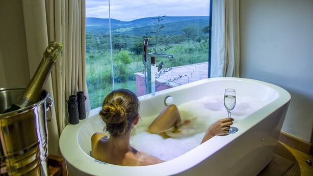 A woman relaxing in a bathtub with a glass of wine, a champagne bottle, and binoculars on a windowsill. - Spirit Of The Masai Mara (Maasai Mara)