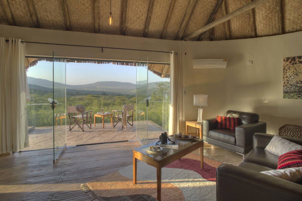 Ein Zimmer mit Glastür, die auf einen Balkon mit schöner Aussicht führt. Im Inneren befindet sich eine Ledersofa, ein Holztisch und ein kleiner Tisch mit einer Lampe.