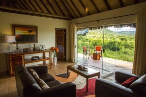 A living room with a sofa, coffee table, and a painting on the wall. There is a sliding glass door leading to an outdoor area with a view of greenery and hills.