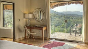A bedroom with a bed, a desk with a chair, a mirror, and a view of a mountainous landscape through a sliding glass door. - Spirit Of The Masai Mara (Maasai Mara)