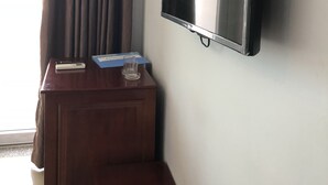 Ein Zimmer mit einem an der Wand befestigten Flachbildfernseher, einem Holztischchen mit Fernbedienung und einem Glas sowie einem Vorhangfenster.