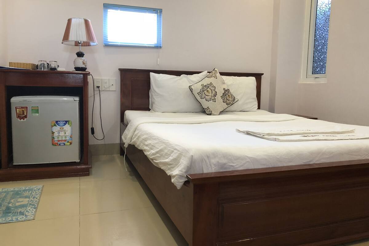 A neatly arranged bedroom with a bed, a small desk with a refrigerator, a lamp, and a window with blinds.